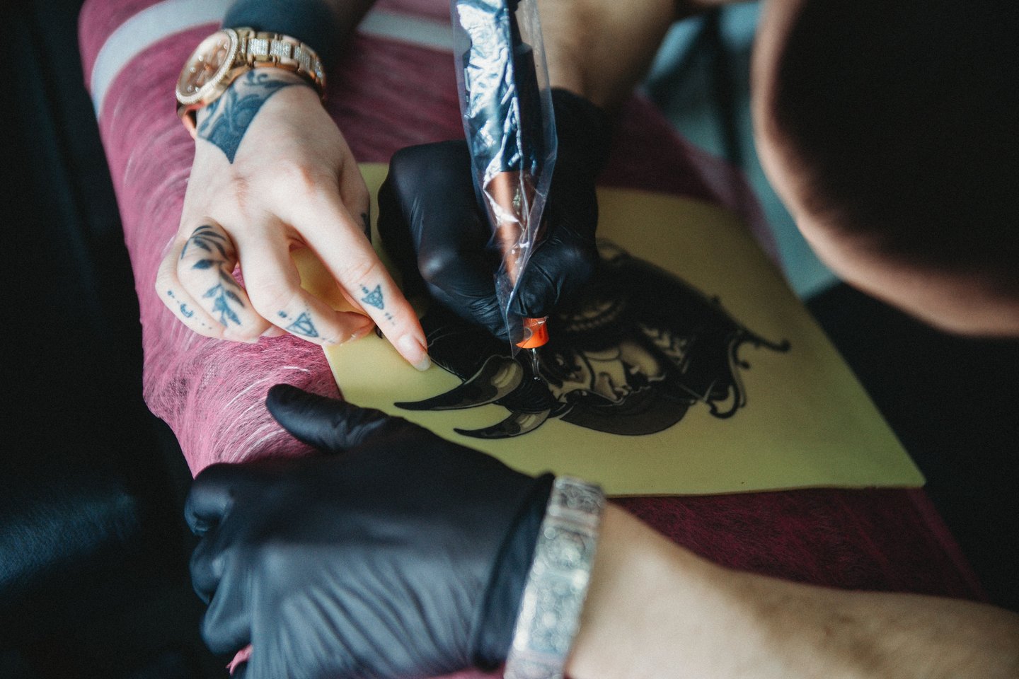 Hands-on tattoo course in Tallinn: mentor guides a student practicing linework on artificial skin with a tattoo machine, both wearing black gloves for hygiene.