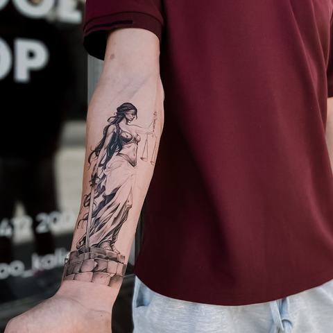 A woman holding scales stretches vertically along the forearm, combining delicate linework, subtle shading and classical symbolism in one composition