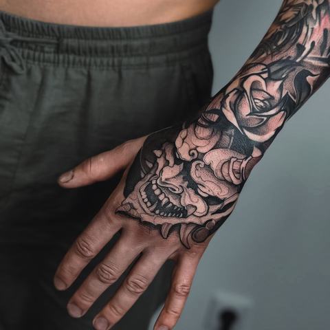 A decorative mask, large roses and dense black shading cover the hand and forearm, forming a powerful composition filled with intricate details